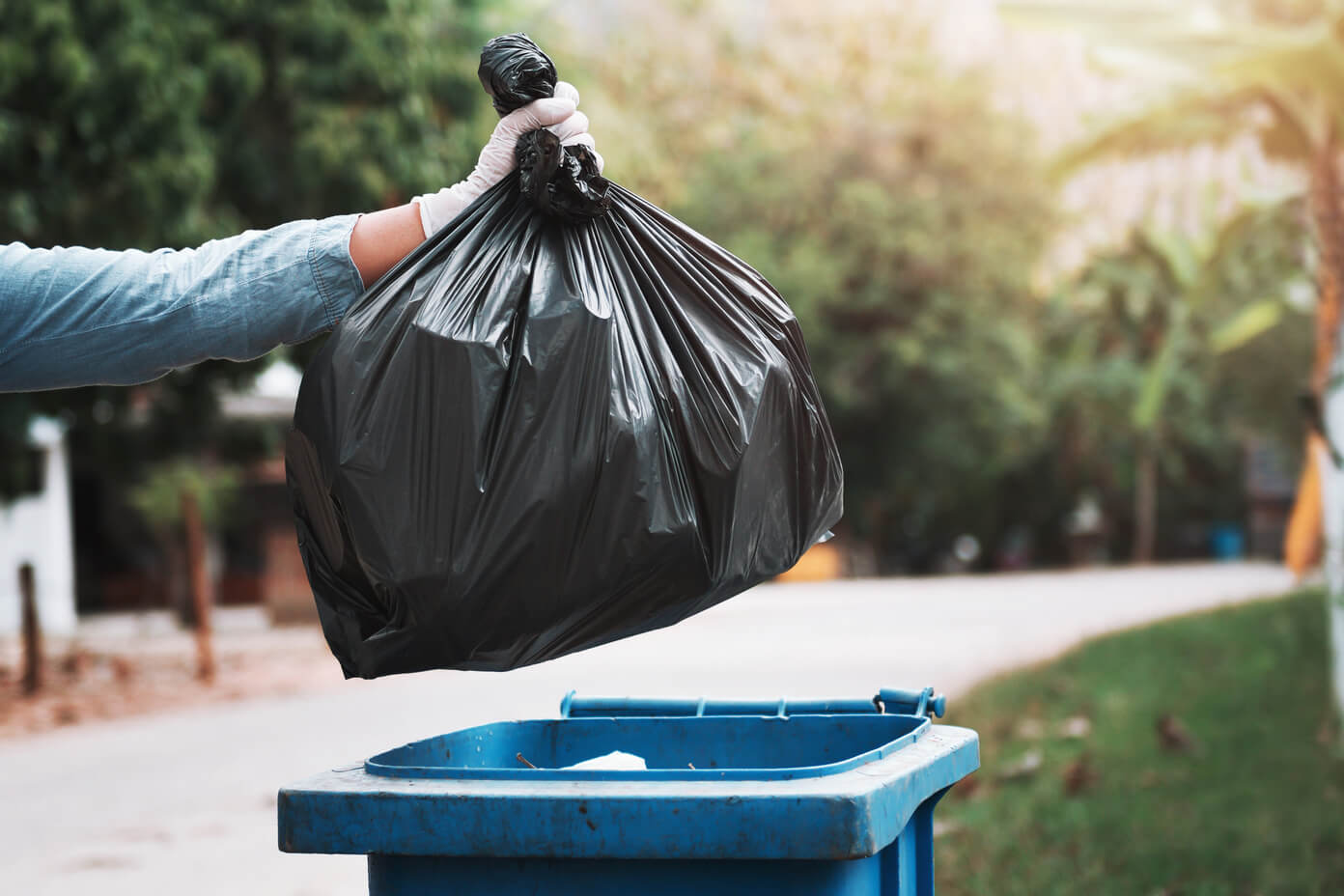 An extended arm holds a full black trash bag over a blue trash bin
